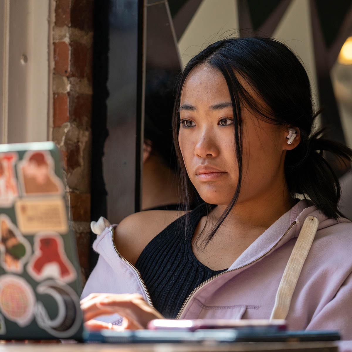 A student wears earbuds and works on her laptop in Cafe Rachel on a sunny day